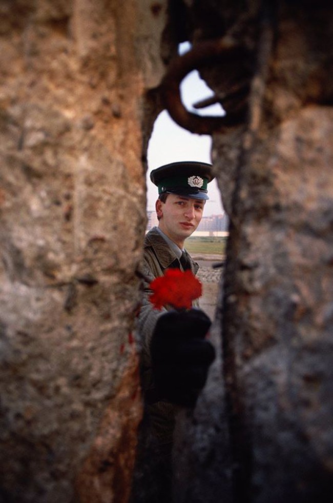 在柏林圍牆倒塌的當天早上，一位東德的首位士兵透過縫隙遞上一朵花。[1989]