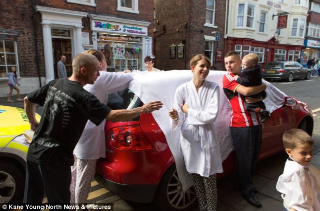 Passersby and members of the wedding party throw hotel towels over the car for privacy