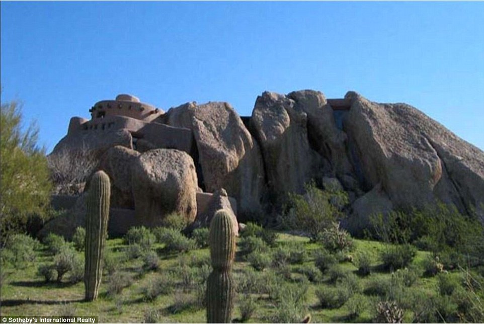The exterior of the home is surrounded by rocks and various desert wildlife, including cacti, trees and small shrubs and bushes